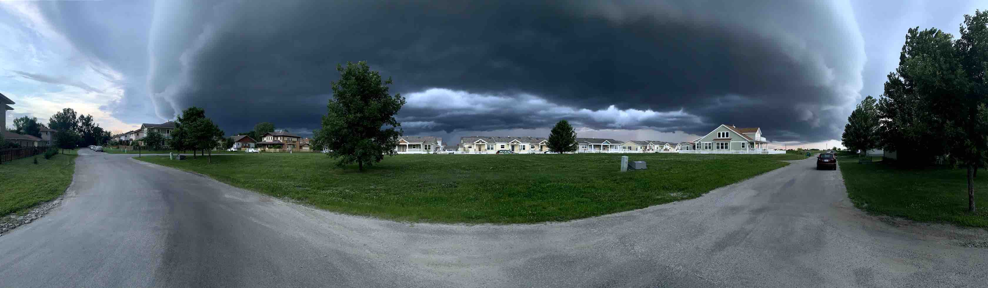 Panoramic image of a neighborhood with houses in the back and a cloudy blue sky with a some trees, taken in the middle of the road